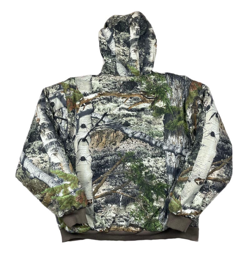 May include: A hooded jacket with a camouflage pattern featuring birch tree bark, green foliage, and brown accents. The jacket has a brown ribbed hem and cuffs. The hood is fully lined with the same camouflage pattern.