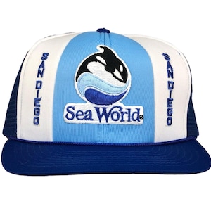 Puede incluir: Gorra de camionero Sea World azul, blanca y negra. La gorra tiene una visera azul y una parte trasera de malla, con las palabras "San Diego" bordadas en los laterales. El panel frontal muestra un logotipo de Sea World y las palabras "Sea World".
