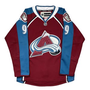 Puede incluir: Una camiseta de hockey granate con detalles azules y blancos. La camiseta presenta el número 9 en las mangas, un logotipo del equipo en el pecho y el logotipo de Reebok. Tiene un cuello blanco y rayas azules en las mangas.