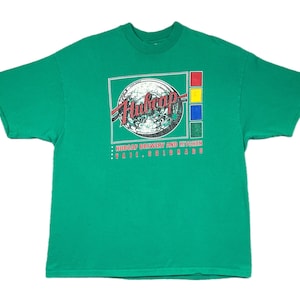 Op de afbeelding: Een groen t-shirt met een grafisch ontwerp. Het ontwerp bevat de tekst "Hubcap Brewery and Kitchen" en "Vail, Colorado". Het ontwerp heeft een cirkelvormig embleem met rode, witte en zwarte accenten en een vierkant met rode, gele, blauwe en groene blokken.