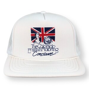 Op de afbeelding: Witte truckercap met een blauwe en rode Union Jack-vlag, een klein beeld van het Verenigd Koninkrijk en de tekst "The Colorado English Motoring Conclave".
