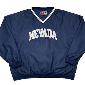 May include: Navy blue pullover windbreaker with a white and gray "NEVADA" arched text across the chest. The jacket has a V-neck collar with white trim and elastic cuffs and hem. The tag reads "2XL".