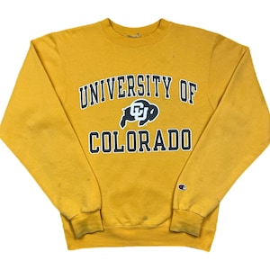 May include: Mustard yellow Champion sweatshirt with the text "UNIVERSITY OF COLORADO" in navy blue arched across the chest. The sweatshirt features a navy blue and white logo of a buffalo. The Champion logo is on the left sleeve.