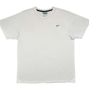 May include: A white t-shirt with a small black Nike swoosh logo on the left chest. The shirt has a crew neck and short sleeves. The tag is visible at the back of the neck.