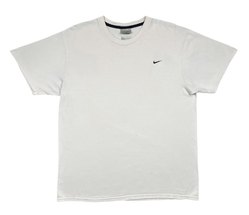 May include: A white t-shirt with a small black Nike swoosh logo on the left chest. The shirt has a crew neck and short sleeves. The tag is visible at the back of the neck.