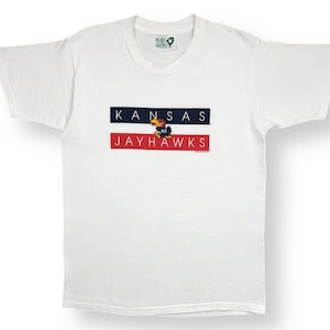 Könnte beinhalten: Weißes T-Shirt mit einem blau-roten Streifenmuster mit dem Text "KANSAS" und "JAYHAWKS" und einem Cartoon-Maskottchen in der Mitte.