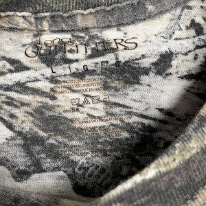 May include: Close-up of a camouflage-patterned t-shirt. The shirt has the text "Lodge Outfitters" printed on it. The shirt also has a tag with washing instructions and fabric content.
