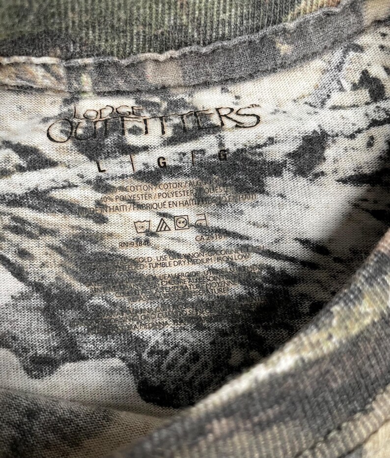 May include: Close-up of a camouflage-patterned t-shirt. The shirt has the text "Lodge Outfitters" printed on it. The shirt also has a tag with washing instructions and fabric content.