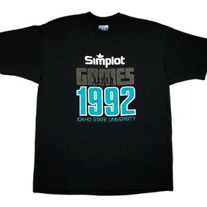 Può includere: Maglietta nera con una grafica blu turchese e bianca che dice "Simplot Games 1992 Idaho State University".