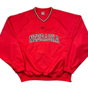 May include: A red Nike pullover windbreaker with a V-neck collar. The word "NEBRASKA" is printed across the chest in white with a black outline. A small black Nike swoosh logo is visible above the text.