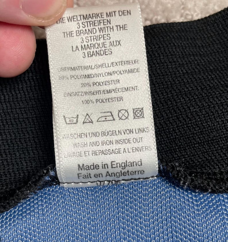 May include: A white tag with black text, including "THE BRAND WITH THE 3 STRIPES" and washing instructions. The tag is attached to a blue and black fabric. The tag also says "Made in England".