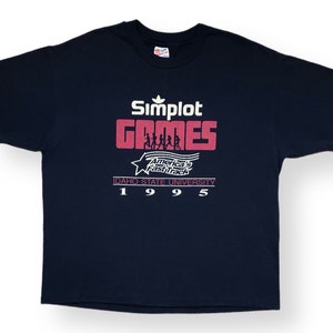 Può includere: Una maglietta blu navy con un logo Adidas bianco e il testo "Simplot Games" in rosso e bianco. I testi "America's Fast Track" e "Idaho State University" sono anch'essi in rosso e bianco. L'anno "1995" è stampato in bianco.