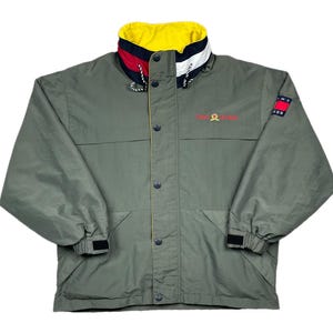 May include: Olive green jacket with a yellow, red, and white collar. The jacket has a button-up front, two pockets, and the Tommy Hilfiger logo embroidered in red. A small Tommy Hilfiger flag logo is on the sleeve.