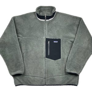 May include: A full-zip, sage green fleece jacket with a black chest pocket. The jacket has a high collar with a white and black trim. The jacket has a small logo on the chest pocket.