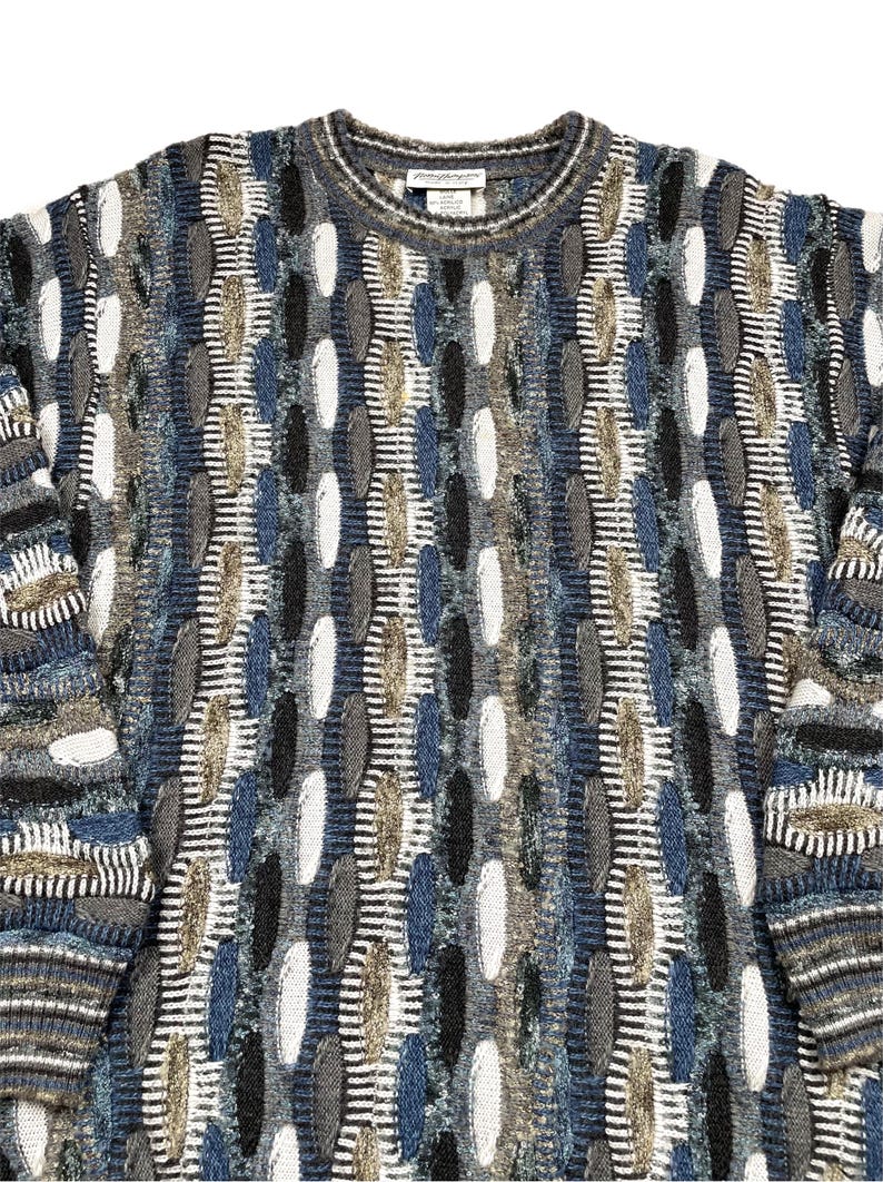 May include: A textured crew neck jumper in shades of blue, grey, white, and tan. The jumper features an all-over pattern of vertical stripes and oval shapes. The sleeves and neckline have a ribbed design. The tag is visible at the back of the neck.