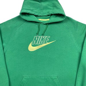 May include: Green Nike hoodie with a yellow Nike logo on the front. The hoodie has a hood with a drawstring and a front pocket. The Nike logo is a swoosh and the word "NIKE" in a sans-serif font.