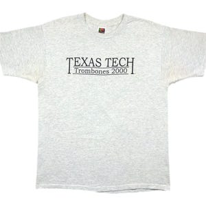 Könnte beinhalten: Hellgraues T-Shirt mit kurzen Ärmeln. Das Shirt trägt den Text "TEXAS TECH" über "Trombones 2000" in schwarzer Schrift. Das Shirt ist aus einem weichen, bequemen Material gefertigt.