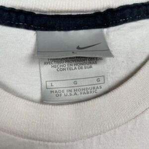 May include: Close-up of a white t-shirt with a black collar. A Nike tag is visible, with text indicating the shirt was made in Honduras of U.S.A. fabric. The tag also includes size information.