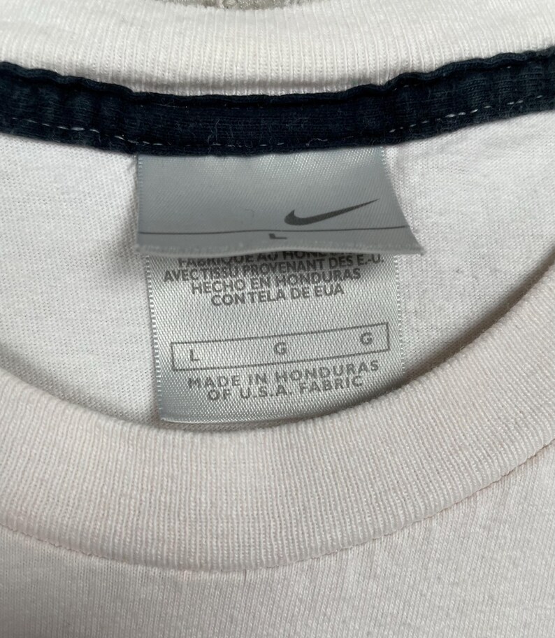 May include: Close-up of a white t-shirt with a black collar. A Nike tag is visible, with text indicating the shirt was made in Honduras of U.S.A. fabric. The tag also includes size information.