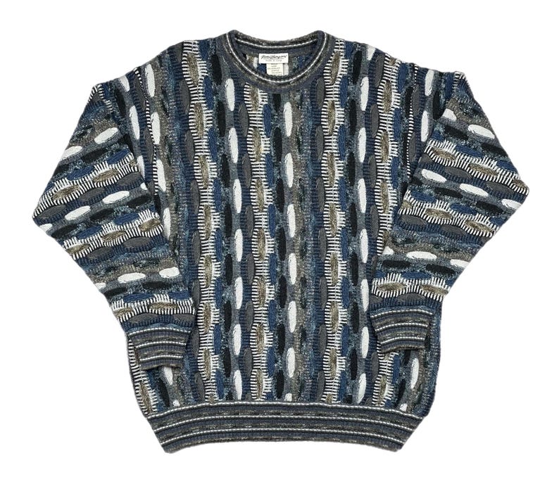 May include: A patterned sweater in shades of blue, grey, white, and brown. The crew neck sweater has a distinctive textured design with long sleeves and ribbed cuffs and hem. The pattern combines geometric and organic shapes, creating a visually interesting garment.