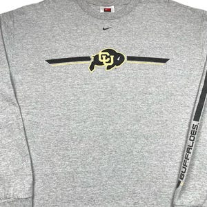 Op de afbeelding: Een witte keramische mok met een zwart handvat en de tekst "But First, Coffee" in een vetgedrukte, zwarte letter. De mok staat tegen een vage achtergrond. De mok is ongeveer 10 cm hoog.