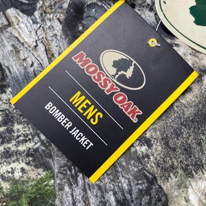 May include: A black tag with yellow trim displays the Mossy Oak logo and the words "MENS BOMBER JACKET". The tag is attached to a camouflage-patterned fabric, with shades of green, brown, and gray.