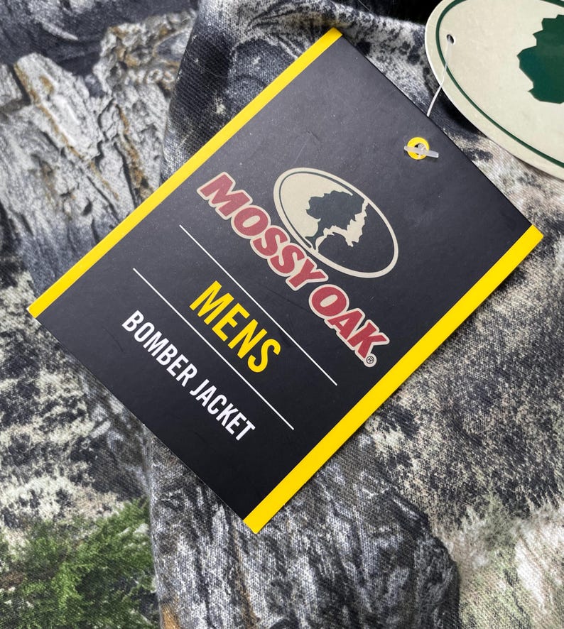 May include: A black tag with yellow trim displays the Mossy Oak logo and the words "MENS BOMBER JACKET". The tag is attached to a camouflage-patterned fabric, with shades of green, brown, and gray.