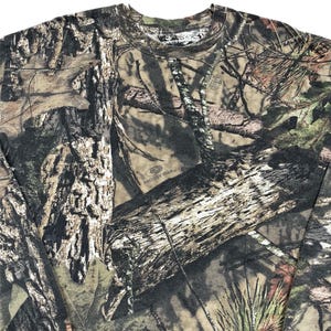 May include: A long-sleeved camouflage shirt featuring a detailed pattern of tree bark, leaves, and branches in shades of brown, green, and tan. The shirt has a crew neck and is made of a soft, comfortable material. The Mossy Oak logo is visible.