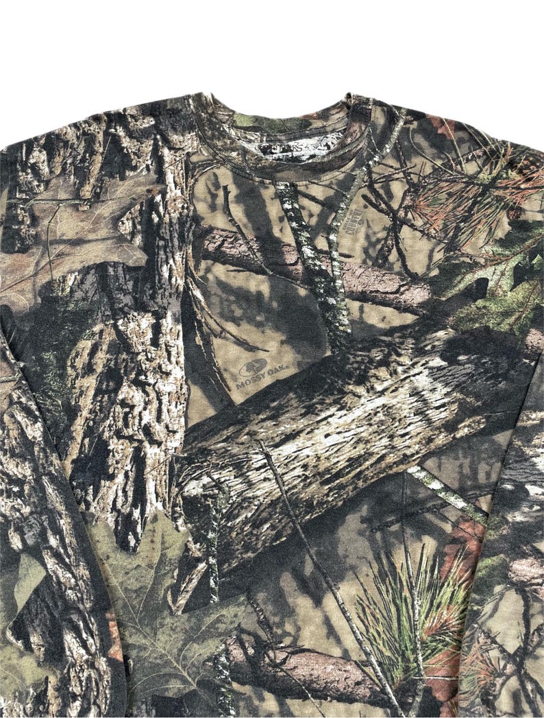 May include: A long-sleeved camouflage shirt featuring a detailed pattern of tree bark, leaves, and branches in shades of brown, green, and tan. The shirt has a crew neck and is made of a soft, comfortable material. The Mossy Oak logo is visible.