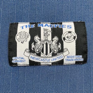 May include: A rectangular black and white patch with the text "THE MAGPIES" in blue. The patch features the Newcastle United crest and the words "OFFICIAL GARMENT" and "REGISTERED TRADE MARKS".