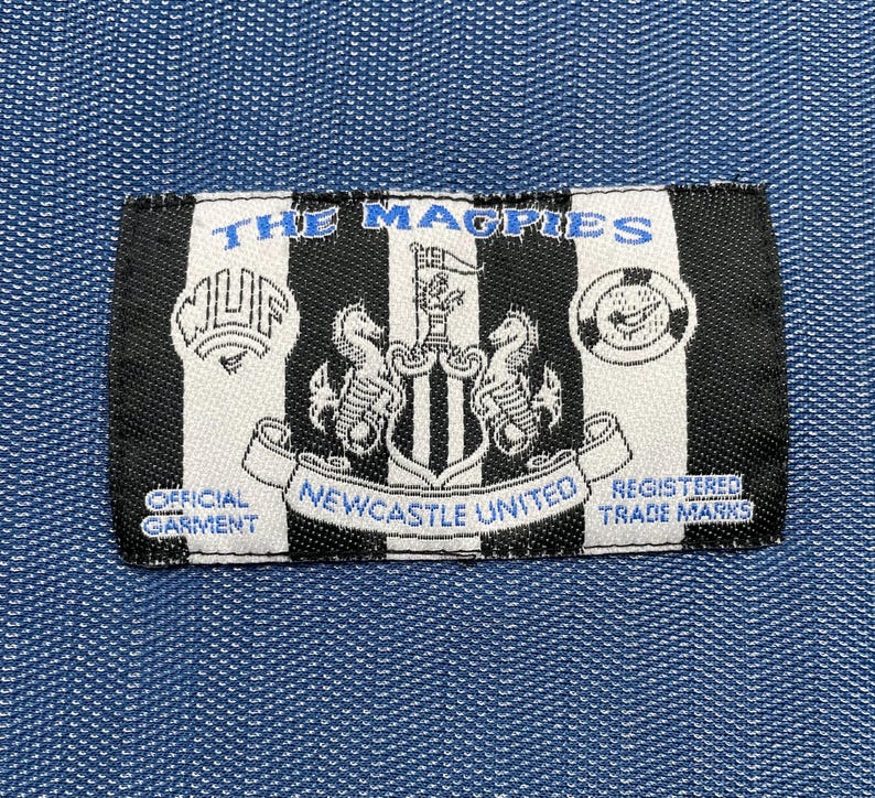 May include: A rectangular black and white patch with the text "THE MAGPIES" in blue. The patch features the Newcastle United crest and the words "OFFICIAL GARMENT" and "REGISTERED TRADE MARKS".