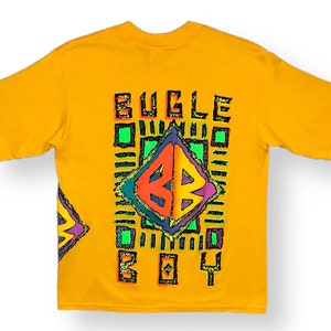 May include: A yellow short-sleeved t-shirt with a colorful graphic print on the back. The graphic features a diamond shape with the letters "BB" inside, surrounded by a geometric pattern and the text "BUGLE BOY".
