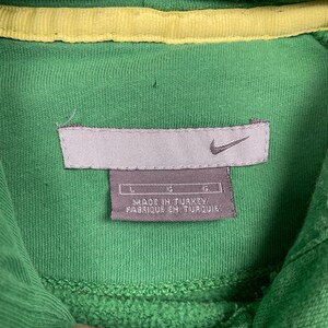 May include: Close-up of a green hooded sweatshirt with a yellow interior lining. The Nike logo is visible on a gray tag, along with the text "MADE IN TURKEY / FABRIQUE EN TURQUIE". The sweatshirt is a size large.