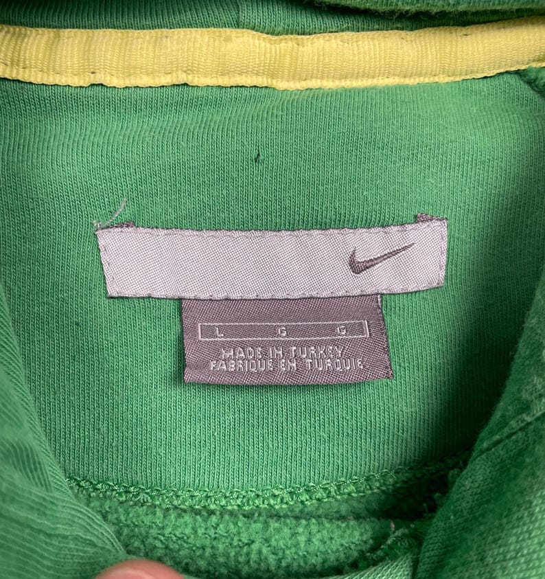May include: Close-up of a green hooded sweatshirt with a yellow interior lining. The Nike logo is visible on a gray tag, along with the text "MADE IN TURKEY / FABRIQUE EN TURQUIE". The sweatshirt is a size large.