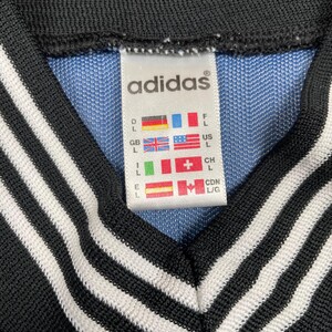 May include: Close-up of an Adidas tag on a blue and black garment. The tag displays the Adidas logo and size information with flags representing different countries. The garment has a black collar with white stripes.