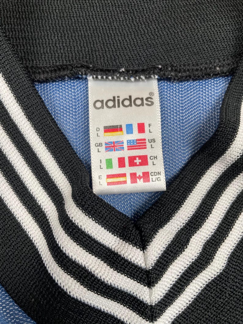 May include: Close-up of an Adidas tag on a blue and black garment. The tag displays the Adidas logo and size information with flags representing different countries. The garment has a black collar with white stripes.