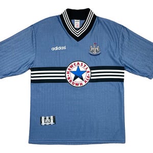 May include: A light blue soccer jersey with short sleeves, featuring a black and white striped design across the chest. The jersey has a V-neck collar with black and white trim, the Adidas logo, and a team crest. The jersey also has the text "NEWCASTLE BROWN ALE".