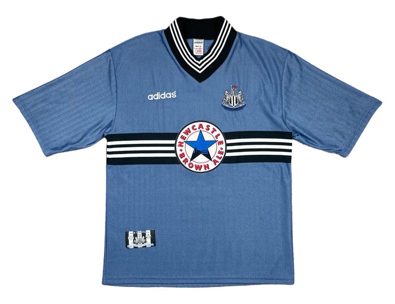 May include: A light blue soccer jersey with short sleeves, featuring a black and white striped design across the chest. The jersey has a V-neck collar with black and white trim, the Adidas logo, and a team crest. The jersey also has the text "NEWCASTLE BROWN ALE".