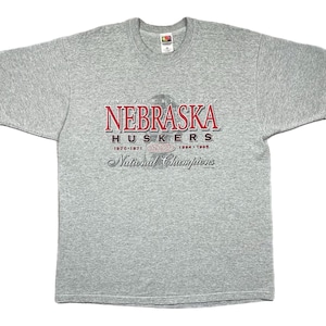 Könnte beinhalten: Graues T-Shirt mit dem Schriftzug "UNIVERSITY OF NEBRASKA HUSKERS" in Rot und Weiß. Das Shirt zeigt auch die Jahre 1970-1971, 1994 und 1995, darunter "National Champions". Klassischer Rundhalsausschnitt.