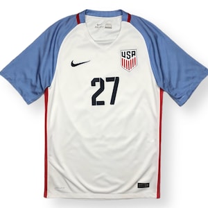 Pode incluir: Camisola de futebol branca com detalhes azuis e vermelhos. A camisola tem o logotipo Nike swoosh, o brasão dos Estados Unidos e o número 27 na frente.