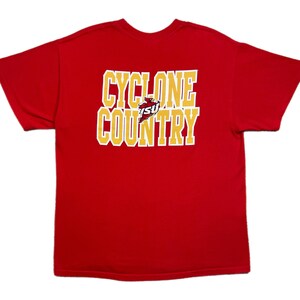 Può includere: Una maglietta rossa con la scritta "CYCLONE COUNTRY" in un design a blocchi dorati e bianchi. La maglietta presenta un logo di un cardinale e le lettere "ISU" al centro del testo.