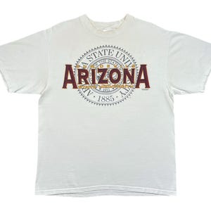 May include: A white short-sleeve t-shirt featuring a large graphic on the chest. The design includes the word ARIZONA in large maroon letters over a circular seal with the text ARIZONA STATE UNIVERSITY 1885 and SUN DEVILS in gold lettering.