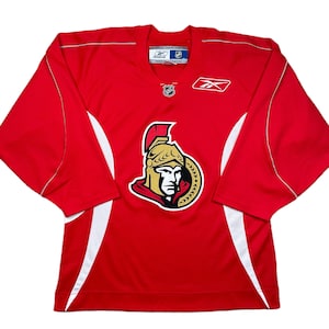 May include: A red hockey jersey with white accents and the Ottawa Senators logo on the front. The jersey features the Reebok logo and NHL emblems. The jersey is a long-sleeved athletic garment.