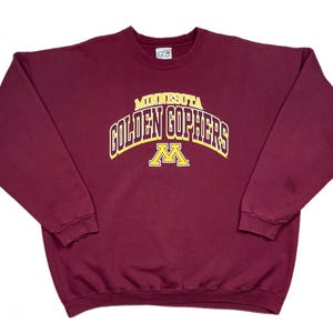 May include: Maroon crew neck sweatshirt featuring the text "MINNESOTA GOLDEN GOPHERS" in gold and white arched lettering. Below the text is a gold "M" logo. The sweatshirt is a classic, long-sleeved design.