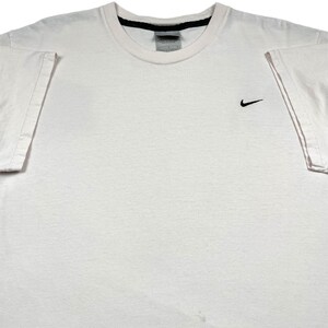 May include: A white crew neck t-shirt with a small black Nike swoosh logo on the left chest. The shirt has short sleeves and a black trim around the neckline. The fabric appears to be cotton.