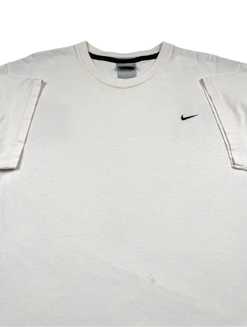 May include: A white crew neck t-shirt with a small black Nike swoosh logo on the left chest. The shirt has short sleeves and a black trim around the neckline. The fabric appears to be cotton.