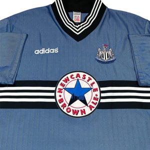 May include: A light blue Adidas Newcastle United soccer jersey. The jersey features a V-neck collar with black and white stripes, the Adidas logo, and the Newcastle United crest. A circular patch reads "Newcastle Brown Ale."