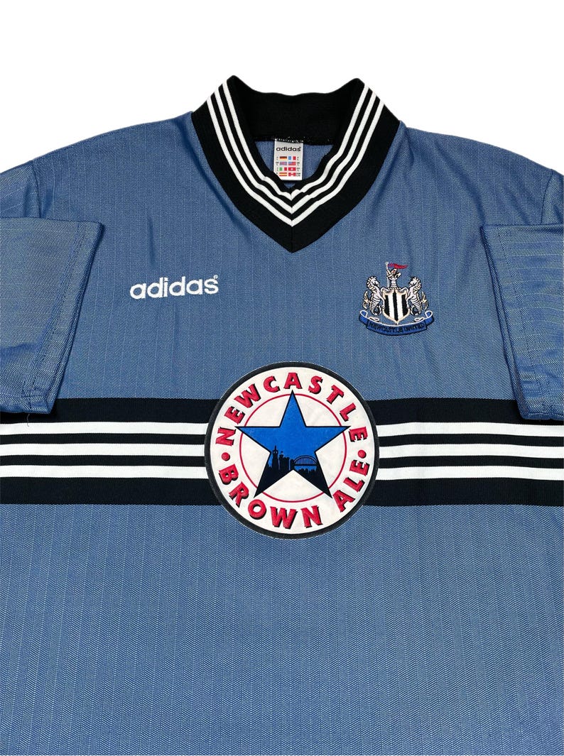 May include: A light blue Adidas Newcastle United soccer jersey. The jersey features a V-neck collar with black and white stripes, the Adidas logo, and the Newcastle United crest. A circular patch reads "Newcastle Brown Ale."