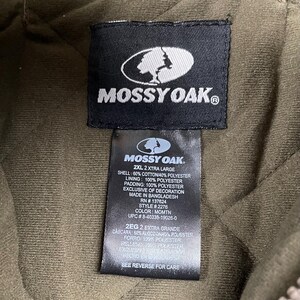 May include: Close-up of a black Mossy Oak label on a quilted olive green jacket. The label includes the Mossy Oak logo, size information, and fabric details. The jacket is 2XL and made in Bangladesh.