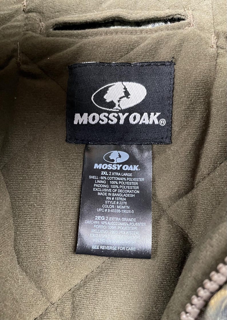 May include: Close-up of a black Mossy Oak label on a quilted olive green jacket. The label includes the Mossy Oak logo, size information, and fabric details. The jacket is 2XL and made in Bangladesh.